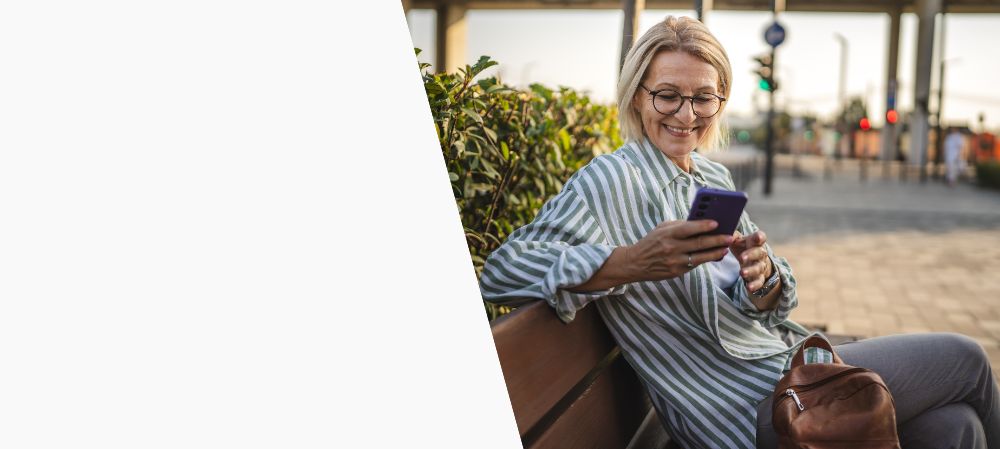 Eine ältere Frau sitzt auf einer Bank und schaut lächelnd auf ihr Handy.